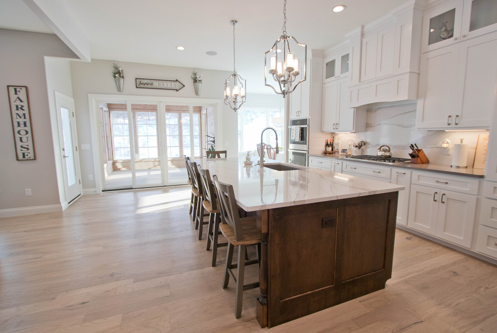Example of a transitional light wood floor and brown floor open concept kitchen design with shaker cabinets, white cabinets, quartz countertops, stone slab backsplash, stainless steel appliances and an island