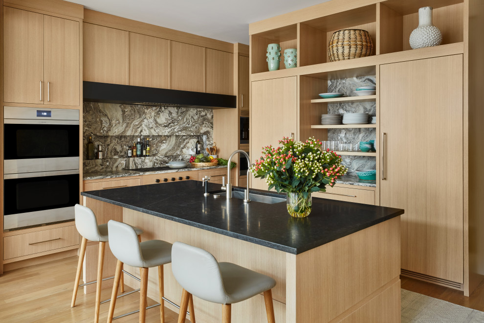 This is an example of a mid-sized transitional l-shaped separate kitchen in Chicago with a drop-in sink, flat-panel cabinets, beige cabinets, marble benchtops, black splashback, marble splashback, panelled appliances, light hardwood floors, with island, beige floor and black benchtop.