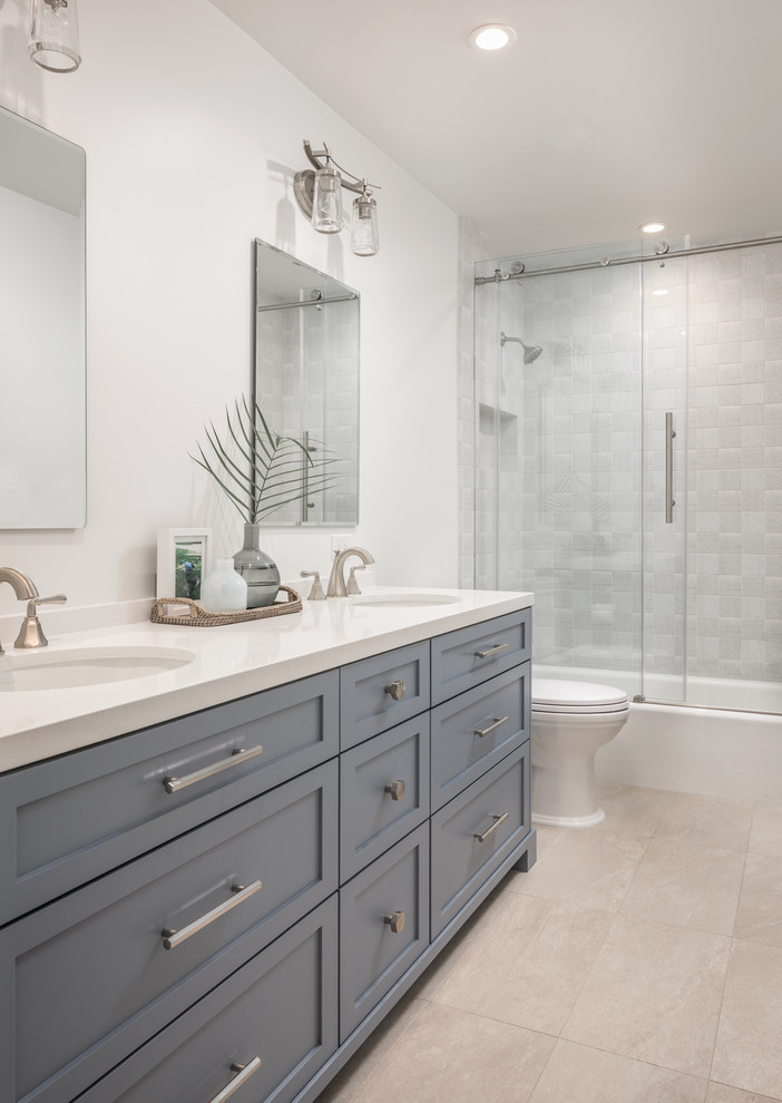 Mid-sized beach style bathroom in Sacramento with shaker cabinets, blue cabinets, a shower/bathtub combo, a one-piece toilet, gray tile, white walls, beige floor and a sliding shower screen.