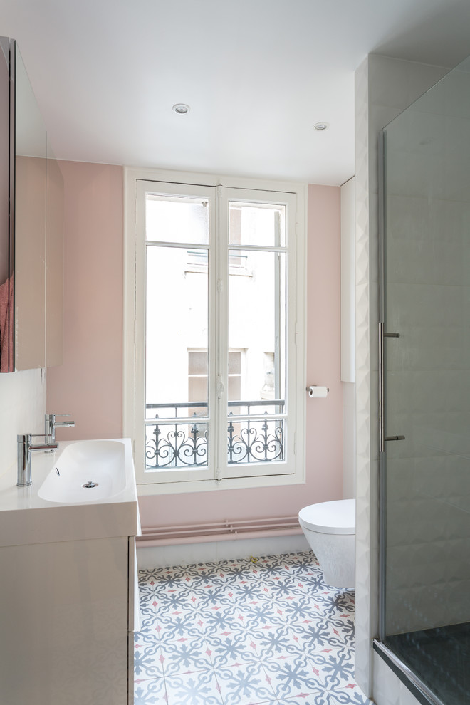 This is an example of a mid-sized contemporary master bathroom in Paris with white cabinets, pink walls, a hinged shower door, flat-panel cabinets, an alcove shower, a wall-mount toilet, white tile, ceramic tile, cement tiles, a trough sink, solid surface benchtops, multi-coloured floor and white benchtops.