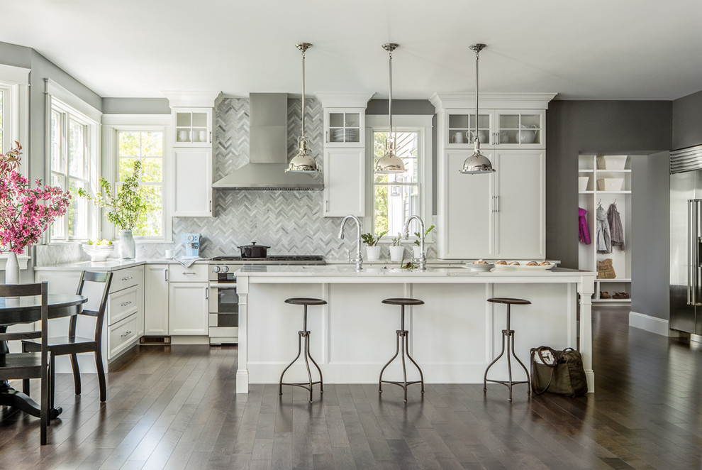 This is an example of a transitional l-shaped eat-in kitchen in Portland Maine with recessed-panel cabinets, white cabinets, marble benchtops, grey splashback, stainless steel appliances, dark hardwood floors and with island.
