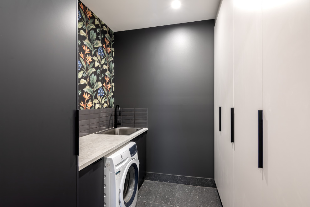 Design ideas for a small modern galley dedicated laundry room in Adelaide with a drop-in sink, flat-panel cabinets, white cabinets, black walls, black floor, black benchtop and wallpaper.