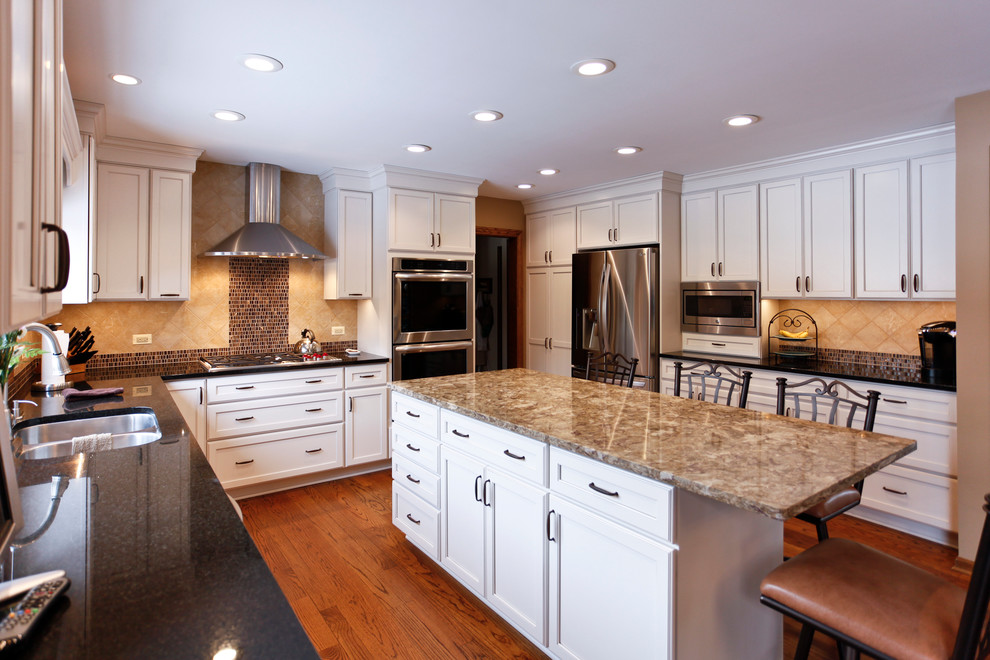Large transitional u-shaped kitchen in Chicago with an undermount sink, recessed-panel cabinets, white cabinets, granite benchtops, beige splashback, ceramic splashback, stainless steel appliances, medium hardwood floors, with island and brown floor.