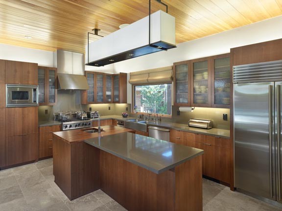 Photo of a mid-sized modern l-shaped kitchen in San Francisco with a farmhouse sink, glass-front cabinets, medium wood cabinets, grey splashback, glass sheet splashback, stainless steel appliances, limestone floors, with island and beige floor.