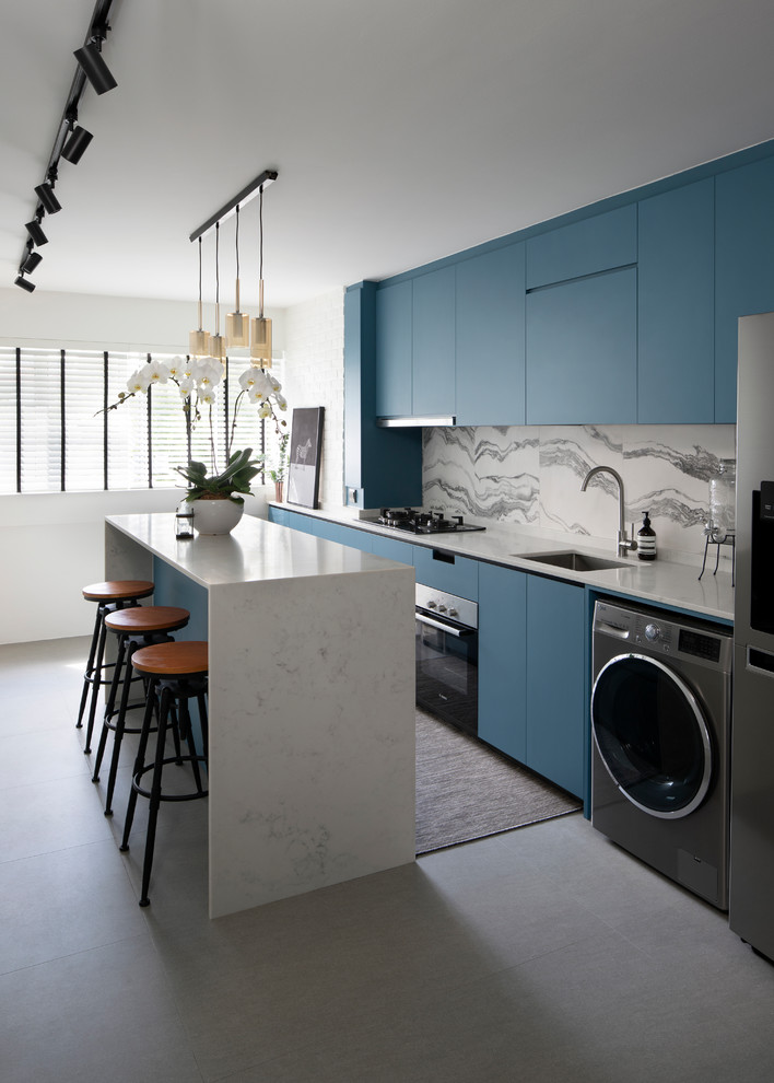 Photo of a contemporary galley kitchen in Singapore with an undermount sink, flat-panel cabinets, blue cabinets, grey splashback, stone slab splashback, stainless steel appliances, porcelain floors, with island, grey floor and white benchtop.