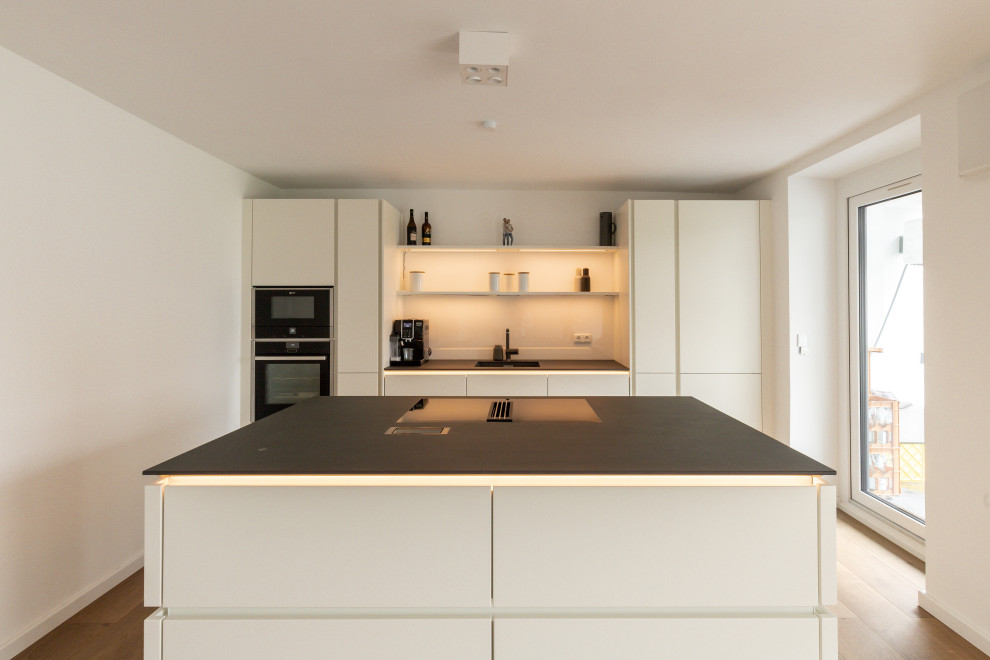 Inspiration for a mid-sized modern galley open plan kitchen in Dusseldorf with an undermount sink, flat-panel cabinets, white cabinets, solid surface benchtops, white splashback, glass sheet splashback, black appliances, light hardwood floors, with island, beige floor and black benchtop.