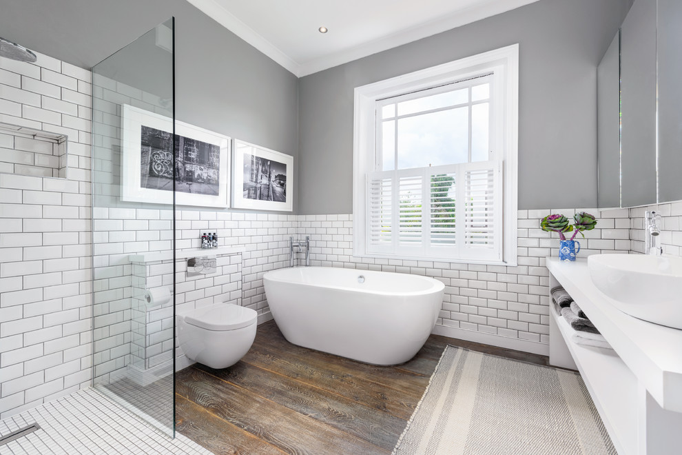 Inspiration for a large modern kids bathroom in London with open cabinets, white cabinets, a freestanding tub, an open shower, a one-piece toilet, white tile, ceramic tile, grey walls, dark hardwood floors, a console sink, solid surface benchtops, brown floor, an open shower and white benchtops.