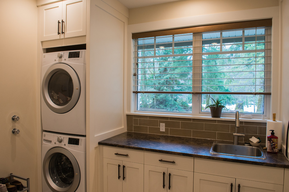 Mid-sized country l-shaped utility room in Vancouver with a single-bowl sink, shaker cabinets, white cabinets, laminate benchtops, beige walls, medium hardwood floors and a stacked washer and dryer.