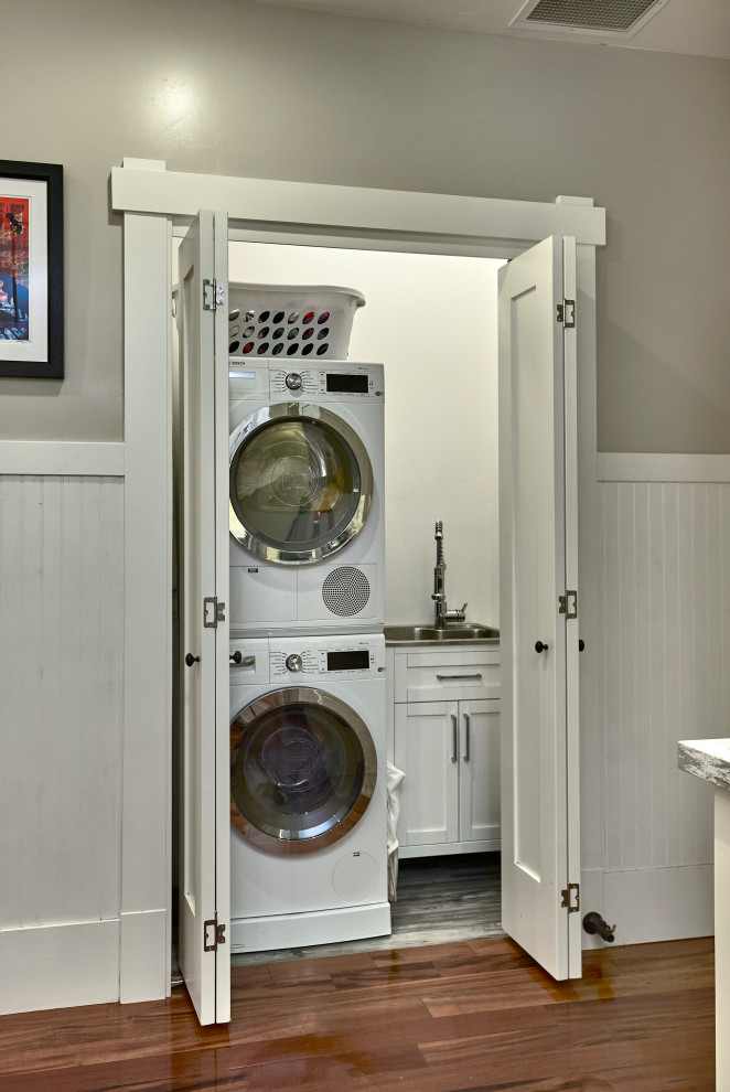 Diseño de armario lavadero lineal de estilo americano pequeño con armarios estilo shaker, puertas de armario blancas, paredes grises, suelo de madera en tonos medios, lavadora y secadora apiladas, suelo rojo, encimeras grises y boiserie