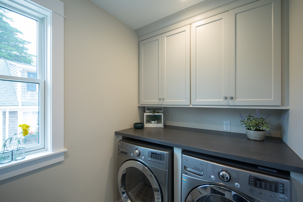 This is an example of a beach style galley dedicated laundry room in Boston with a farmhouse sink, shaker cabinets, white cabinets, solid surface benchtops, beige walls, ceramic floors, a side-by-side washer and dryer, multi-coloured floor and grey benchtop.