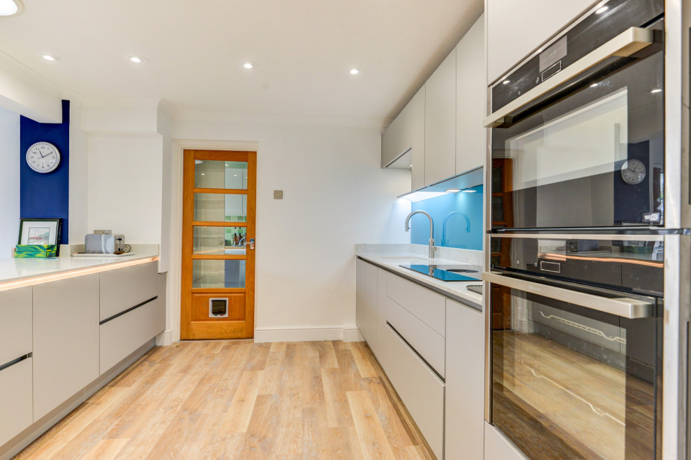 Inspiration for a mid-sized modern single-wall eat-in kitchen in Sussex with an undermount sink, flat-panel cabinets, grey cabinets, quartzite benchtops, blue splashback, glass sheet splashback, panelled appliances, vinyl floors, with island, brown floor and white benchtop.