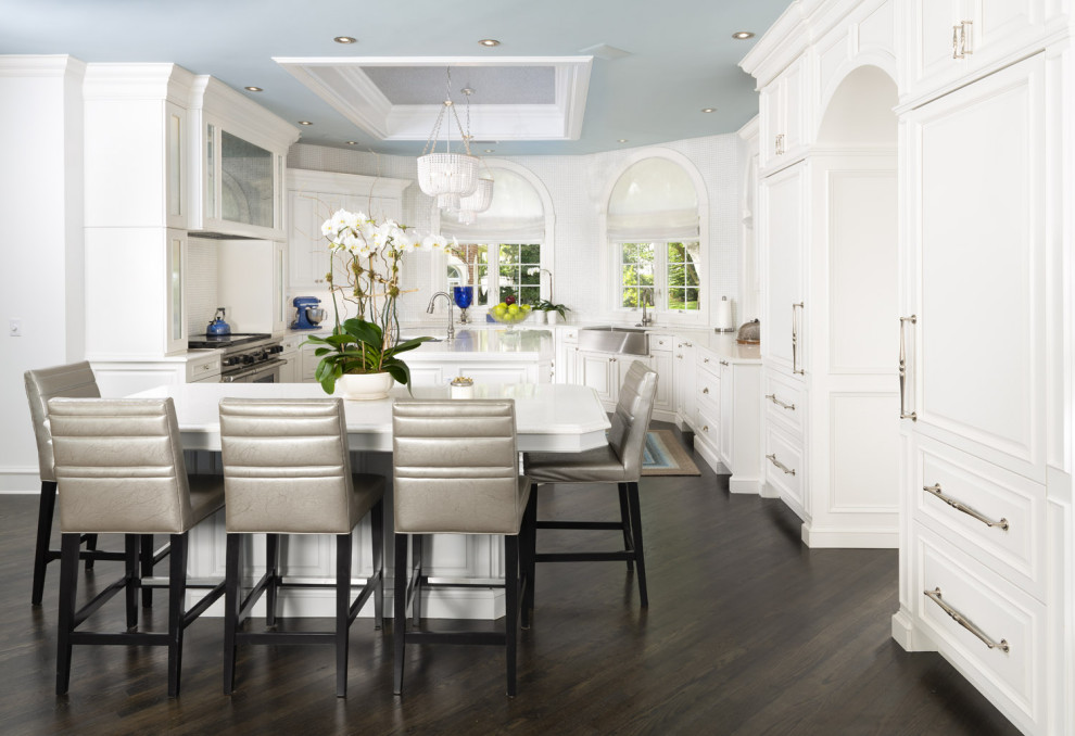 This is an example of a large transitional open plan kitchen in Tampa with a farmhouse sink, raised-panel cabinets, white cabinets, quartz benchtops, white splashback, mosaic tile splashback, panelled appliances, dark hardwood floors, multiple islands, brown floor, white benchtop and wallpaper.