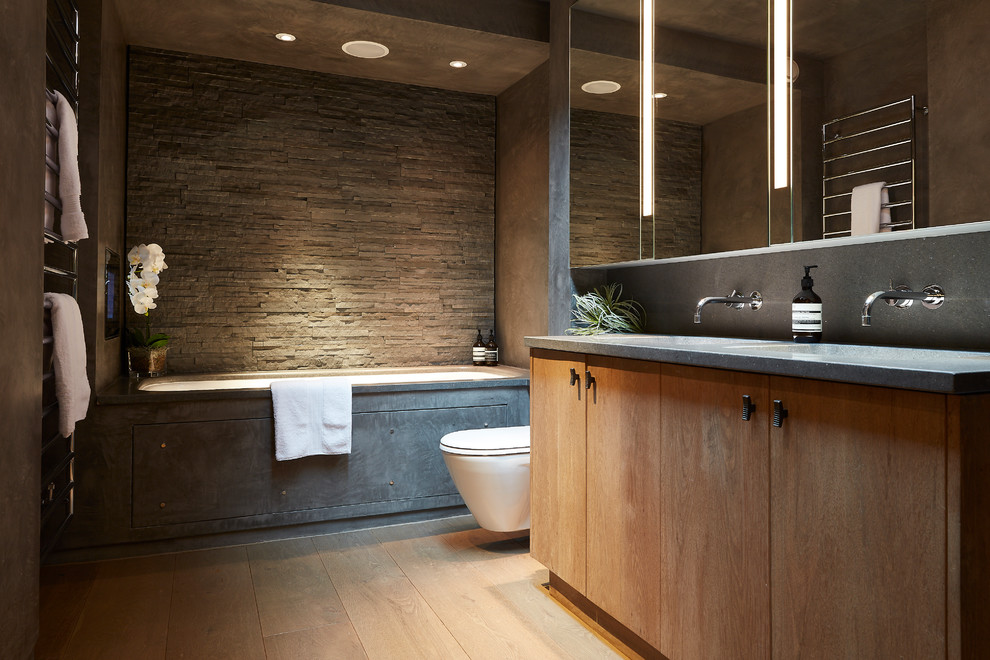 Inspiration for a mid-sized contemporary bathroom in London with flat-panel cabinets, light wood cabinets, a wall-mount toilet, gray tile, grey walls, light hardwood floors, beige floor, grey benchtops, an undermount tub, an integrated sink and concrete benchtops.
