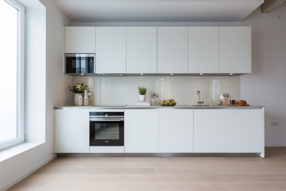 This is an example of a contemporary single-wall kitchen in Gloucestershire with a drop-in sink, flat-panel cabinets, white cabinets, white splashback, glass sheet splashback, black appliances, light hardwood floors, no island, beige floor and grey benchtop.