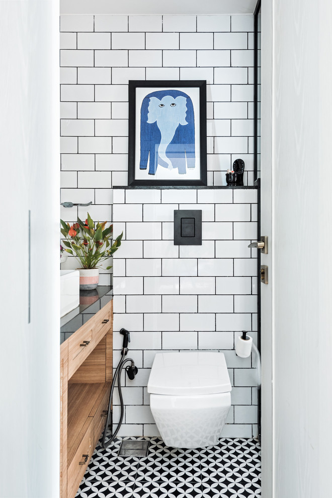 Inspiration for a contemporary bathroom in Mumbai with recessed-panel cabinets, medium wood cabinets, a wall-mount toilet, white tile, subway tile, white walls, a vessel sink and black floor.