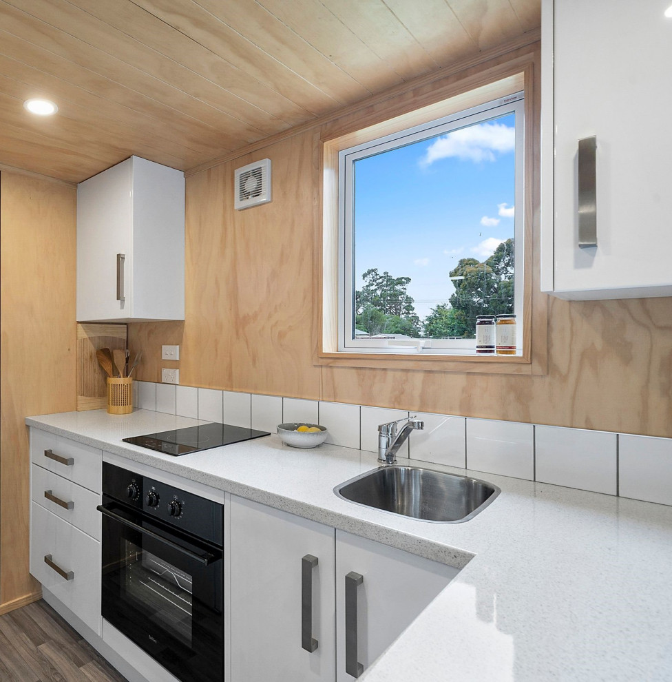 Photo of a small modern l-shaped eat-in kitchen in Wellington with a single-bowl sink, flat-panel cabinets, white cabinets, white splashback, ceramic splashback, black appliances, linoleum floors, brown floor, multi-coloured benchtop and timber.