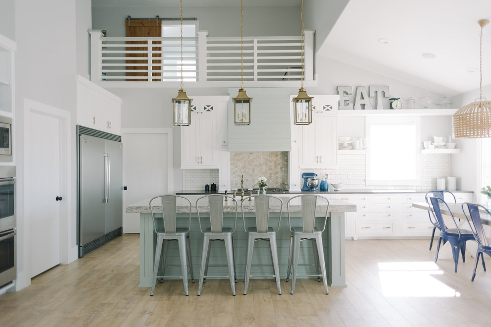 Photo of a large beach style eat-in kitchen in Salt Lake City with with island, shaker cabinets, white cabinets, granite benchtops, white splashback, porcelain splashback, stainless steel appliances, a farmhouse sink and light hardwood floors.