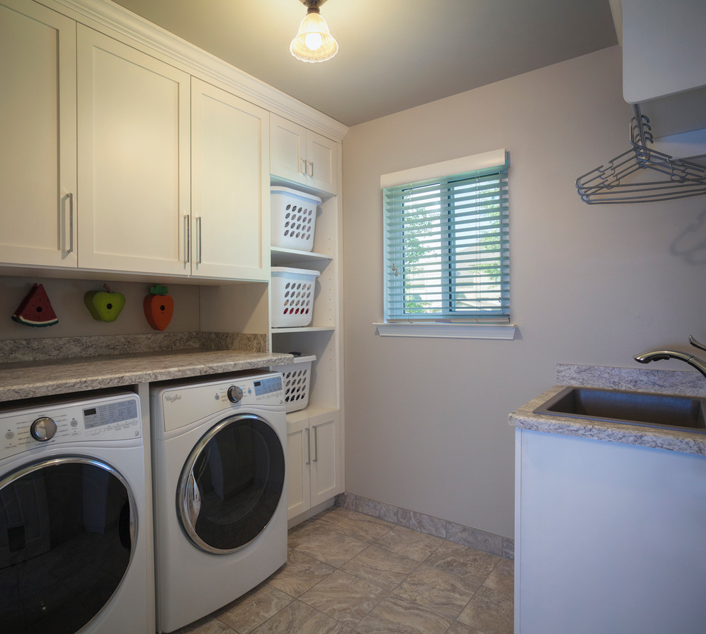 This is an example of a small contemporary galley dedicated laundry room in Detroit with a farmhouse sink, white cabinets, granite benchtops, beige walls, a side-by-side washer and dryer, beige floor, ceramic floors and recessed-panel cabinets.