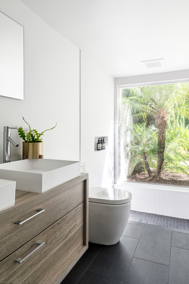Photo of a mid-sized modern master bathroom in Orange County with flat-panel cabinets, light wood cabinets, a curbless shower, a one-piece toilet, white tile, porcelain tile, white walls, porcelain floors, a vessel sink, wood benchtops, black floor, an open shower and white benchtops.