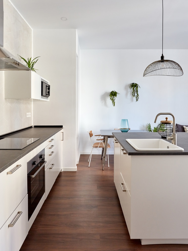 Mid-sized contemporary galley open plan kitchen in Madrid with flat-panel cabinets, white cabinets, concrete benchtops, with island, grey benchtop, a drop-in sink, grey splashback, cement tile splashback, black appliances, dark hardwood floors and brown floor.
