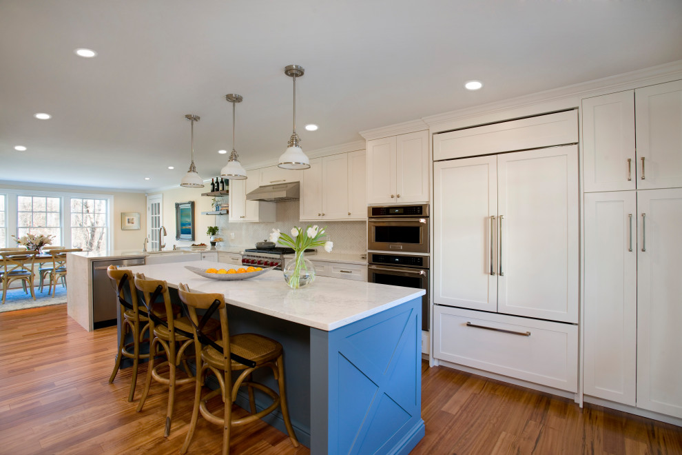 Photo of a large transitional l-shaped kitchen in Boston with a farmhouse sink, shaker cabinets, white cabinets, white splashback, stainless steel appliances, with island, brown floor and white benchtop.