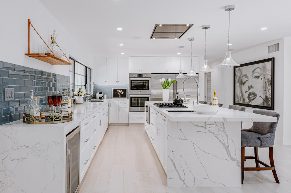 Inspiration for a large beach style l-shaped open plan kitchen in Los Angeles with an undermount sink, shaker cabinets, white cabinets, marble benchtops, blue splashback, stainless steel appliances, light hardwood floors, with island and white benchtop.