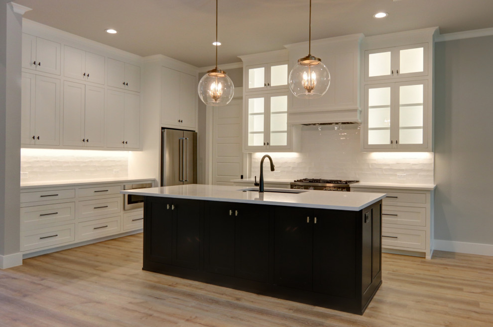 Photo of a large transitional l-shaped kitchen in Dallas with a single-bowl sink, flat-panel cabinets, white cabinets, quartz benchtops, white splashback, ceramic splashback, stainless steel appliances, vinyl floors, with island, brown floor and white benchtop.