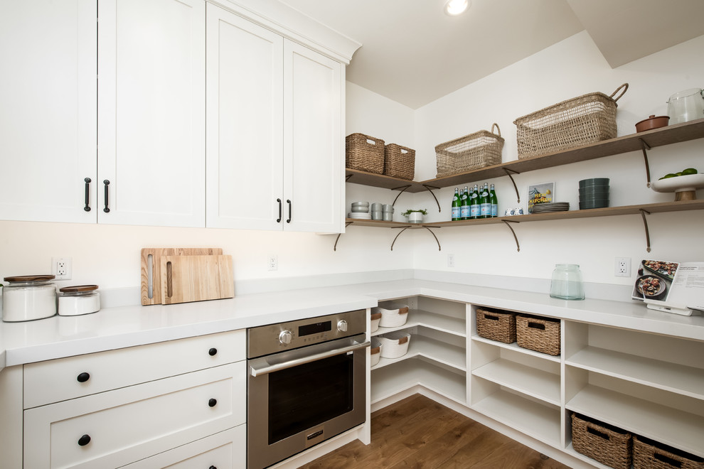 Inspiration for a country l-shaped kitchen pantry in Salt Lake City with shaker cabinets, white cabinets, white splashback, stainless steel appliances, medium hardwood floors, no island, brown floor and white benchtop.