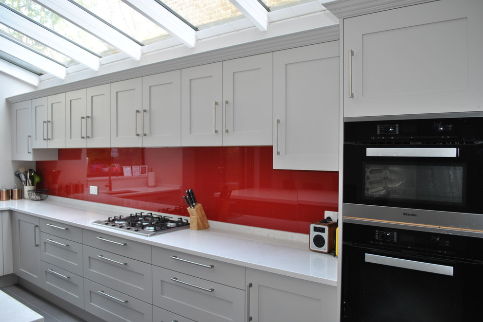 Design ideas for a large modern l-shaped eat-in kitchen in London with a single-bowl sink, shaker cabinets, grey cabinets, quartzite benchtops, red splashback, glass sheet splashback, black appliances, porcelain floors, with island, grey floor and white benchtop.
