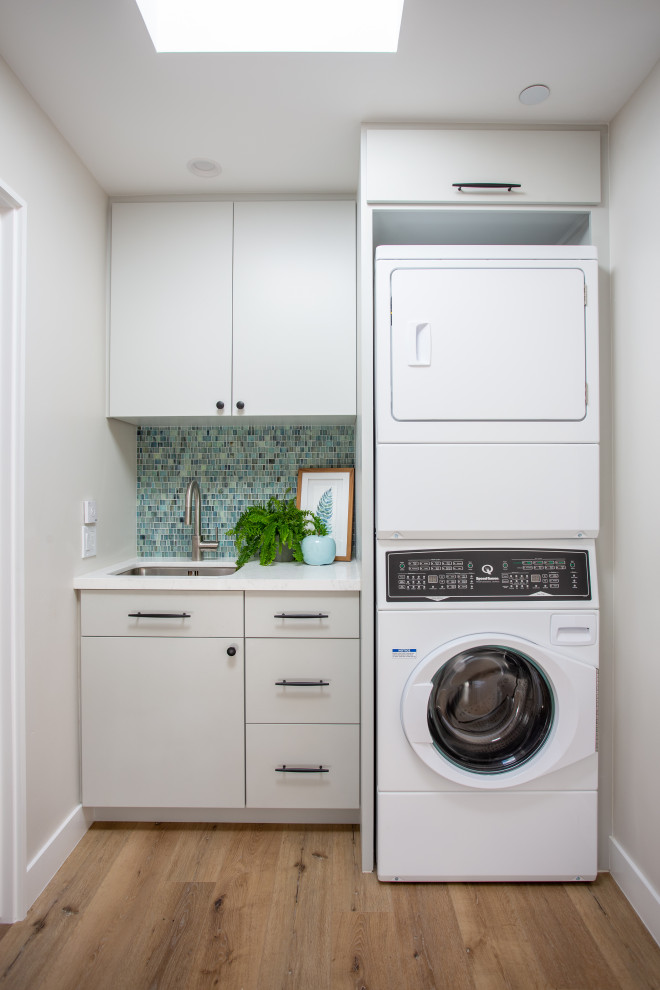 Design ideas for a mid-sized beach style single-wall dedicated laundry room in Santa Barbara with an undermount sink, flat-panel cabinets, grey cabinets, quartz benchtops, blue splashback, glass tile splashback, white walls, vinyl floors, a stacked washer and dryer, brown floor and white benchtop.
