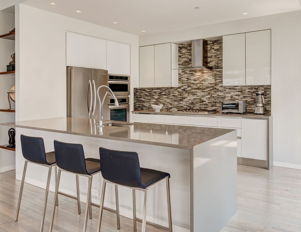 This is an example of a contemporary u-shaped open plan kitchen in New York with an undermount sink, flat-panel cabinets, stainless steel benchtops, multi-coloured splashback, mosaic tile splashback, stainless steel appliances, light hardwood floors, a peninsula, white cabinets, beige floor and grey benchtop.