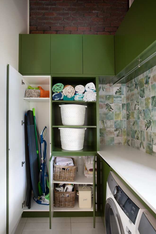 Modern inredning av en liten vita l-formad vitt tvättstuga, med en nedsänkt diskho, gröna skåp, bänkskiva i kvarts, grönt stänkskydd, stänkskydd i porslinskakel, flerfärgade väggar, klinkergolv i porslin, en tvättmaskin och torktumlare bredvid varandra och grått golv
