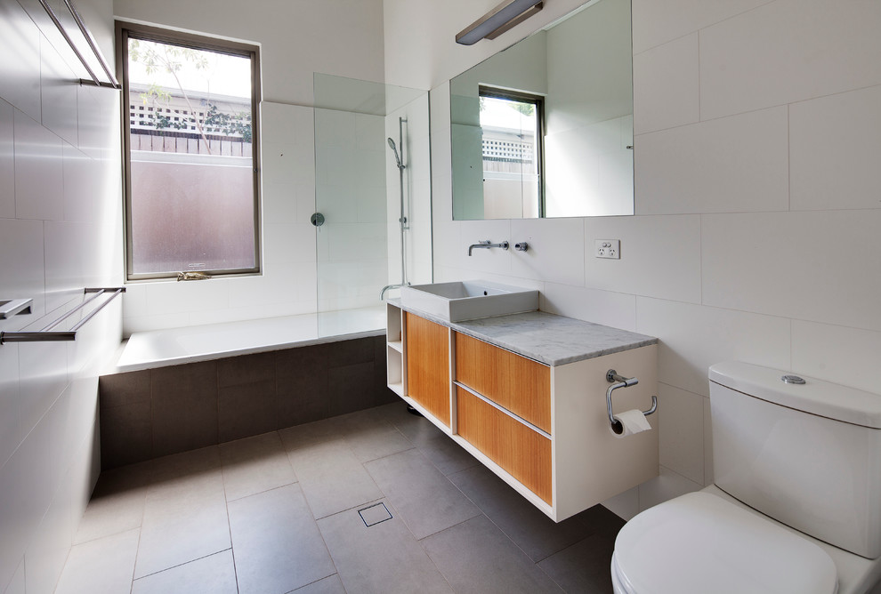 This is an example of a mid-sized contemporary bathroom in Perth with a vessel sink, marble benchtops, a drop-in tub, a two-piece toilet, white tile, white walls, porcelain floors, flat-panel cabinets, medium wood cabinets and a shower/bathtub combo.