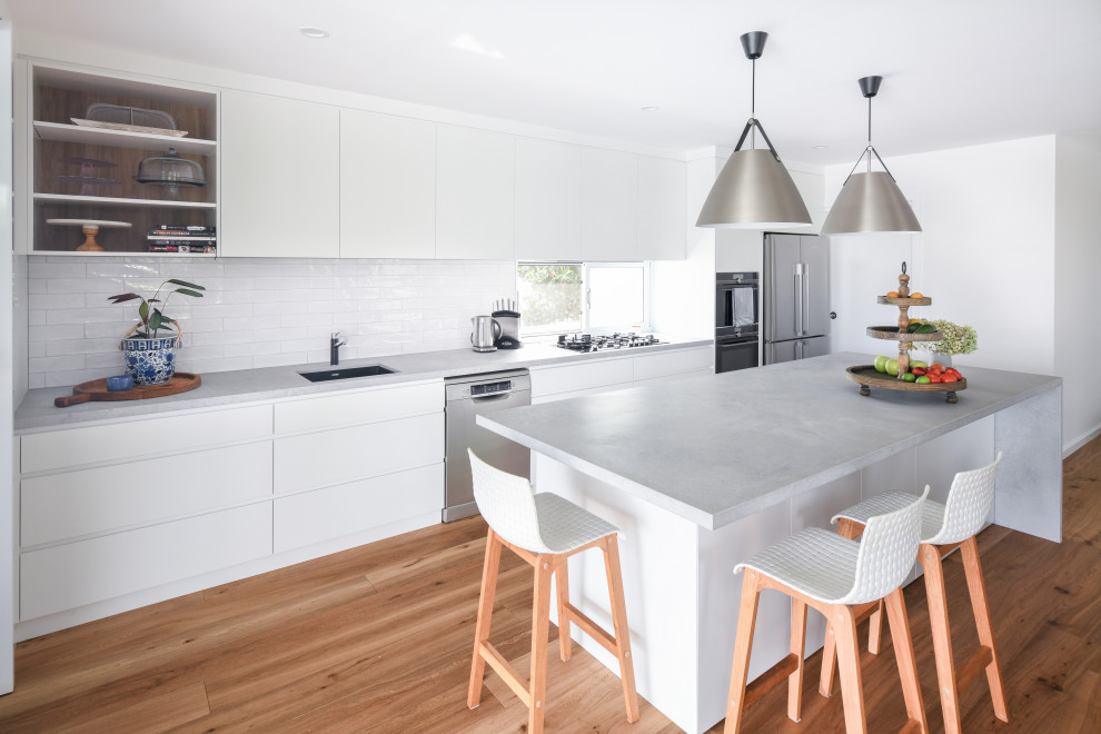 Photo of a mid-sized contemporary single-wall open plan kitchen in Sydney with an undermount sink, flat-panel cabinets, white cabinets, quartz benchtops, white splashback, ceramic splashback, stainless steel appliances, medium hardwood floors, with island, brown floor and grey benchtop.