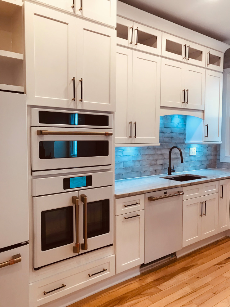 Photo of a large traditional u-shaped kitchen in DC Metro with a single-bowl sink, shaker cabinets, white cabinets, quartzite benchtops, blue splashback, ceramic splashback, white appliances, light hardwood floors, with island, brown floor, white benchtop and exposed beam.