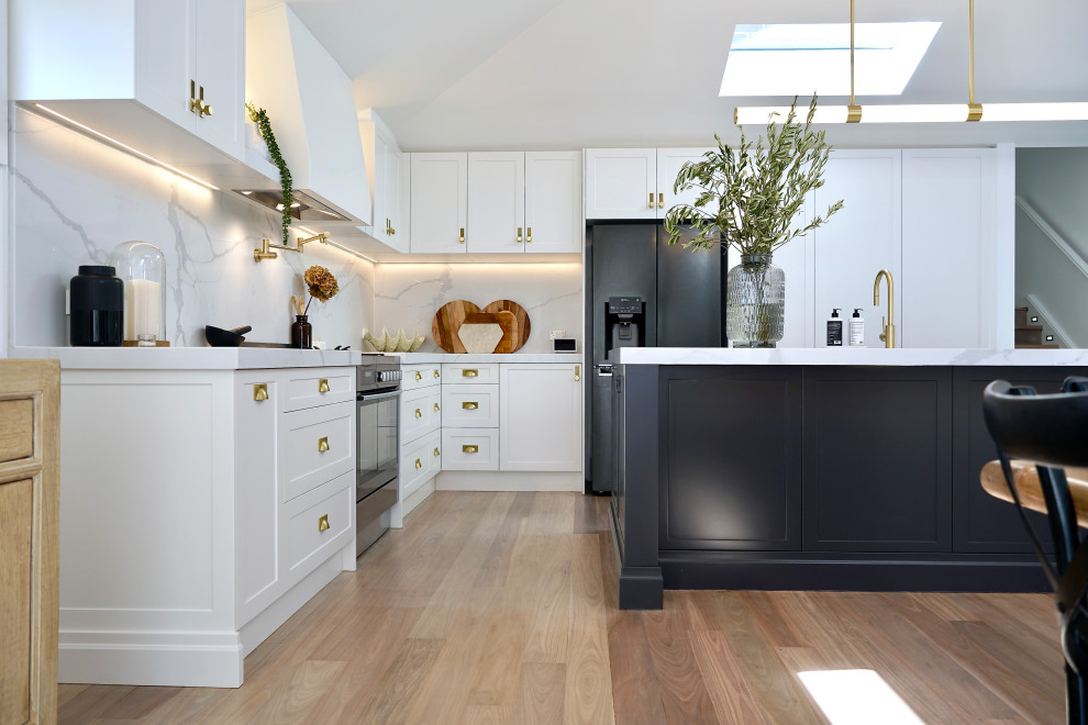 Photo of a large traditional l-shaped open plan kitchen in Sydney with a farmhouse sink, shaker cabinets, white cabinets, quartz benchtops, white splashback, engineered quartz splashback, stainless steel appliances, light hardwood floors, with island, brown floor and white benchtop.
