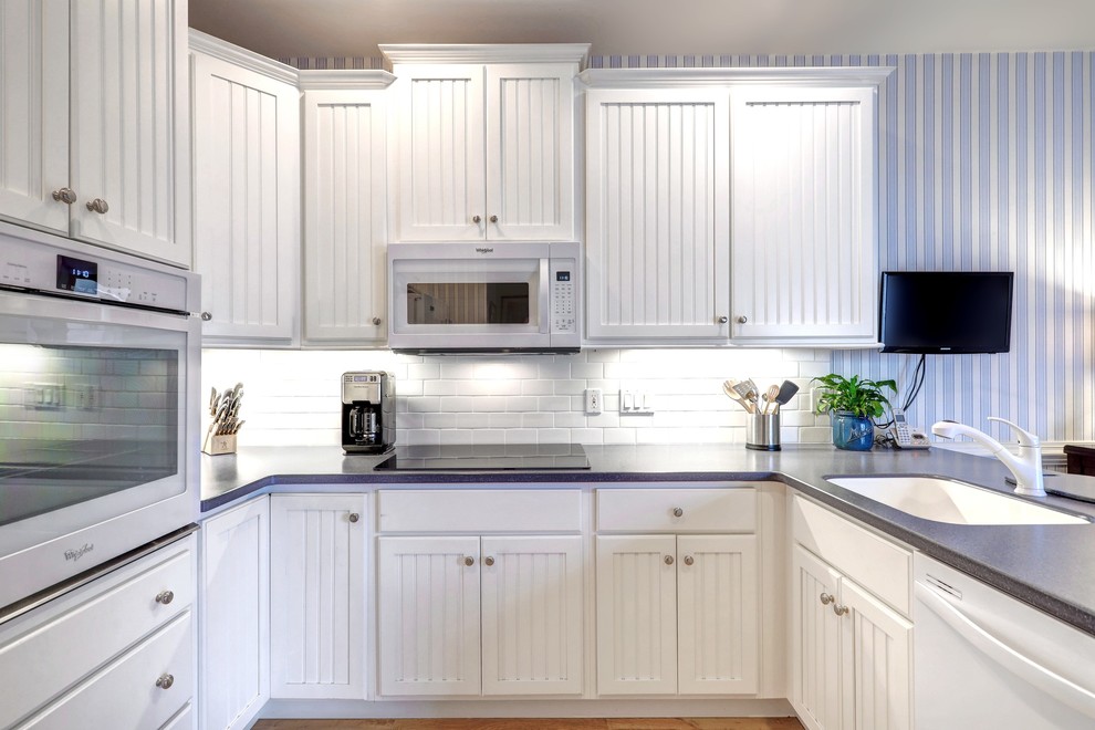 Design ideas for a mid-sized beach style u-shaped kitchen pantry in Other with a single-bowl sink, white cabinets, quartz benchtops, white splashback, subway tile splashback, stainless steel appliances, plywood floors, a peninsula, beige floor and raised-panel cabinets.