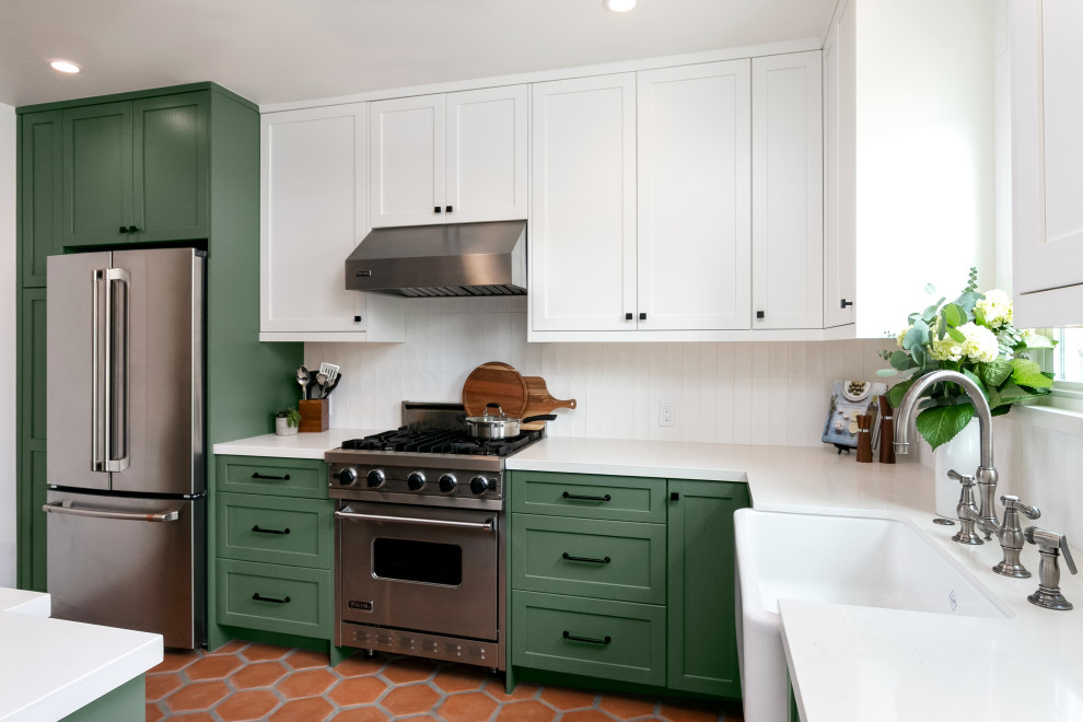 Inspiration for a mid-sized transitional l-shaped terra-cotta tile and brown floor enclosed kitchen remodel in Los Angeles with a farmhouse sink, shaker cabinets, green cabinets, quartz countertops, white backsplash, ceramic backsplash, stainless steel appliances, no island and white countertops