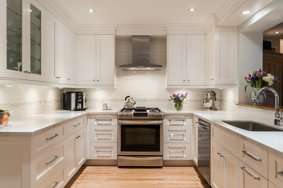 Example of a large transitional u-shaped light wood floor enclosed kitchen design in Toronto with an undermount sink, shaker cabinets, white cabinets, quartz countertops, white backsplash, stone tile backsplash, stainless steel appliances and no island