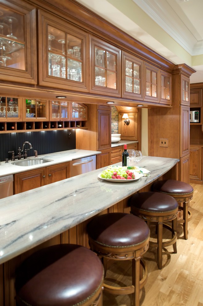 This is an example of a large traditional u-shaped seated home bar in Philadelphia with an undermount sink, glass-front cabinets, medium wood cabinets, marble benchtops, black splashback and light hardwood floors.