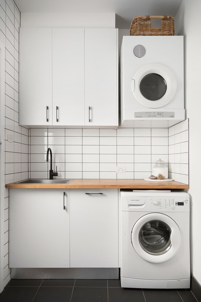 Inspiration for a contemporary single-wall dedicated laundry room in Melbourne with a drop-in sink, flat-panel cabinets, white cabinets, wood benchtops, white walls, black floor and brown benchtop.