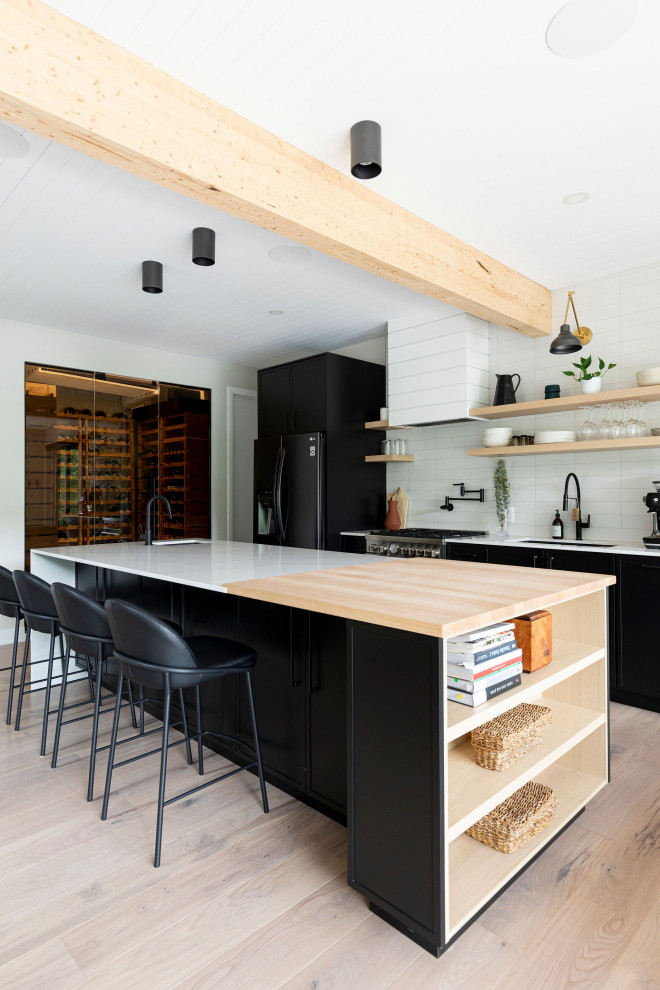 This is an example of a mid-sized country galley open plan kitchen in Montreal with an undermount sink, shaker cabinets, black cabinets, quartz benchtops, white splashback, porcelain splashback, black appliances, light hardwood floors, with island, white benchtop and timber.