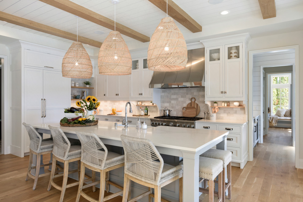 Photo of a beach style l-shaped kitchen in Minneapolis with a farmhouse sink, shaker cabinets, white cabinets, grey splashback, stainless steel appliances, medium hardwood floors, with island, brown floor and white benchtop.