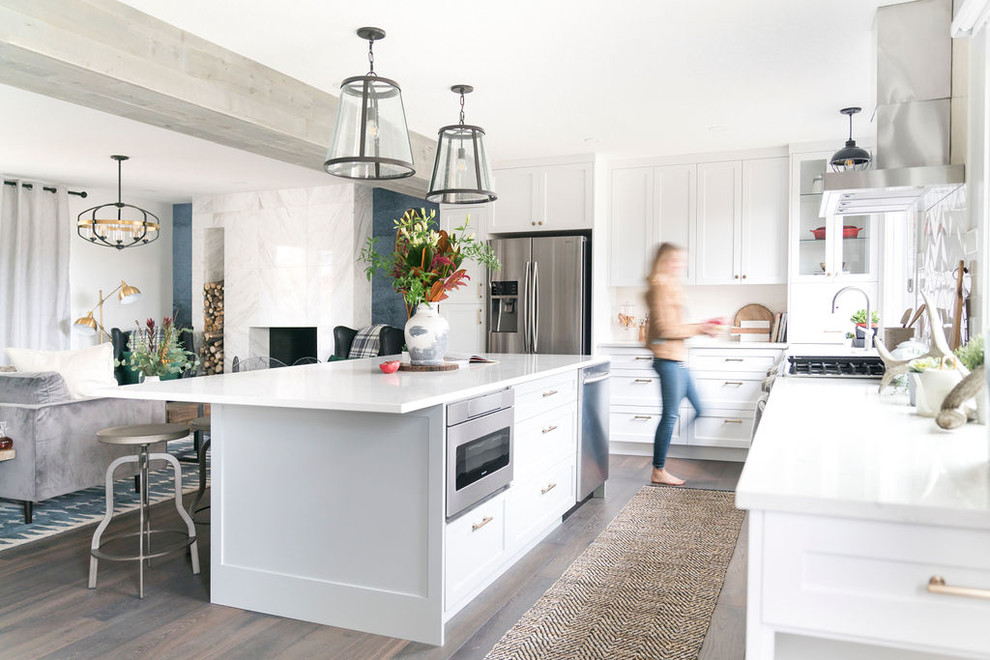 This is an example of a mid-sized transitional l-shaped open plan kitchen in Calgary with a farmhouse sink, shaker cabinets, white cabinets, quartz benchtops, white splashback, stainless steel appliances, dark hardwood floors, with island, brown floor and white benchtop.