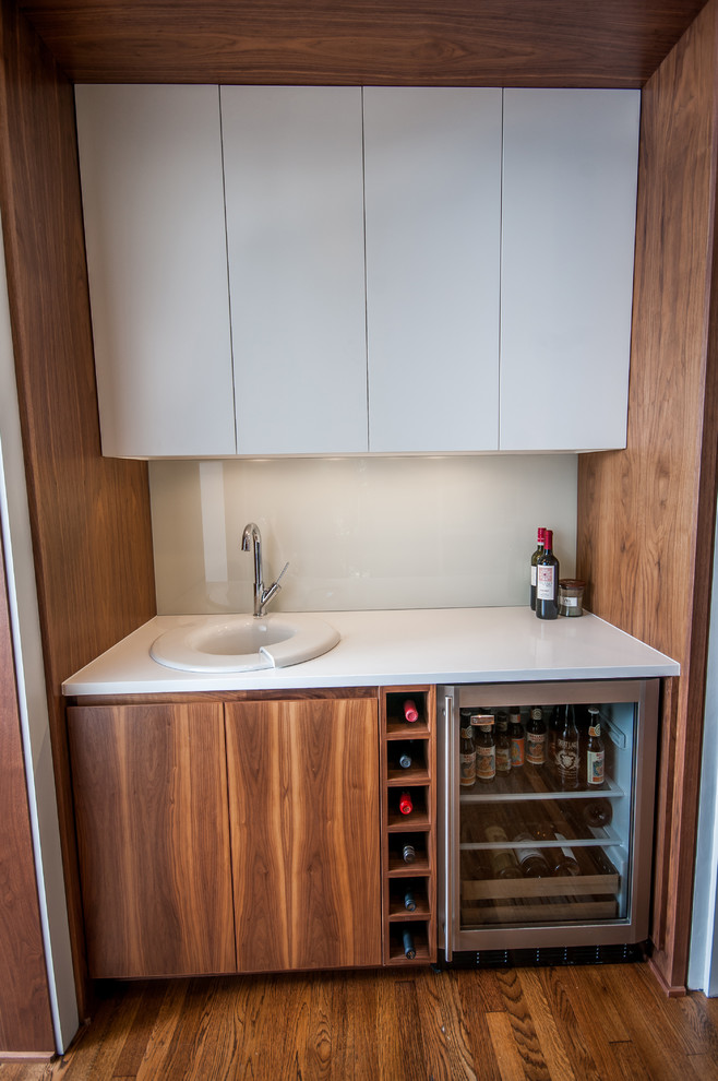 Photo of a small contemporary single-wall wet bar in Cleveland with a drop-in sink, flat-panel cabinets, medium wood cabinets, solid surface benchtops, white splashback, glass sheet splashback, medium hardwood floors and brown floor.