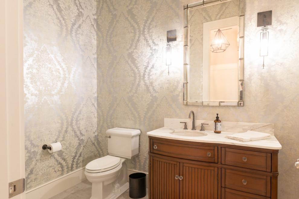 Photo of a country powder room in Houston with furniture-like cabinets, medium wood cabinets, a two-piece toilet, porcelain floors, an undermount sink, marble benchtops, white floor, white benchtops, a built-in vanity and wallpaper.
