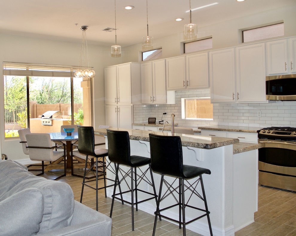 Inspiration for a mid-sized beach style l-shaped open plan kitchen in Los Angeles with a double-bowl sink, raised-panel cabinets, white cabinets, granite benchtops, white splashback, subway tile splashback, stainless steel appliances, porcelain floors, with island and beige floor.