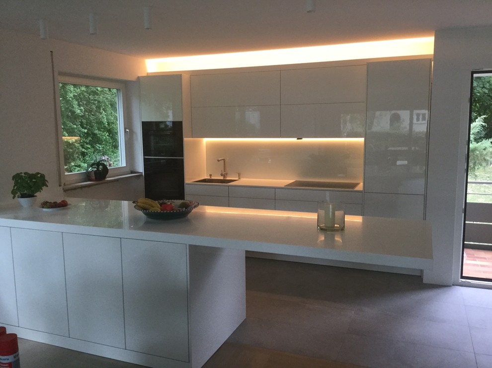 Inspiration for a mid-sized contemporary single-wall open plan kitchen in Other with an undermount sink, flat-panel cabinets, white cabinets, solid surface benchtops, white splashback, glass sheet splashback, black appliances, with island and ceramic floors.
