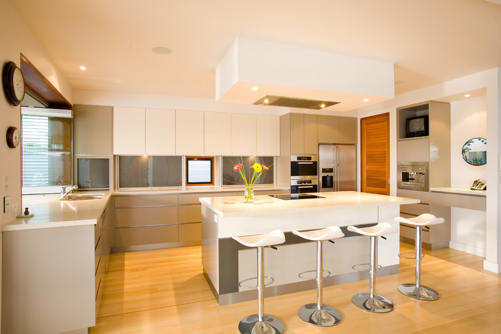 This is an example of a large contemporary l-shaped kitchen in Sunshine Coast with flat-panel cabinets, stainless steel appliances, light hardwood floors, white benchtop, an undermount sink, grey splashback, glass sheet splashback, with island, beige floor and white cabinets.