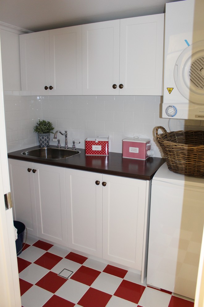 Inspiration for a small beach style single-wall dedicated laundry room in Brisbane with a drop-in sink, shaker cabinets, white cabinets, laminate benchtops, white walls, a stacked washer and dryer and red floor.